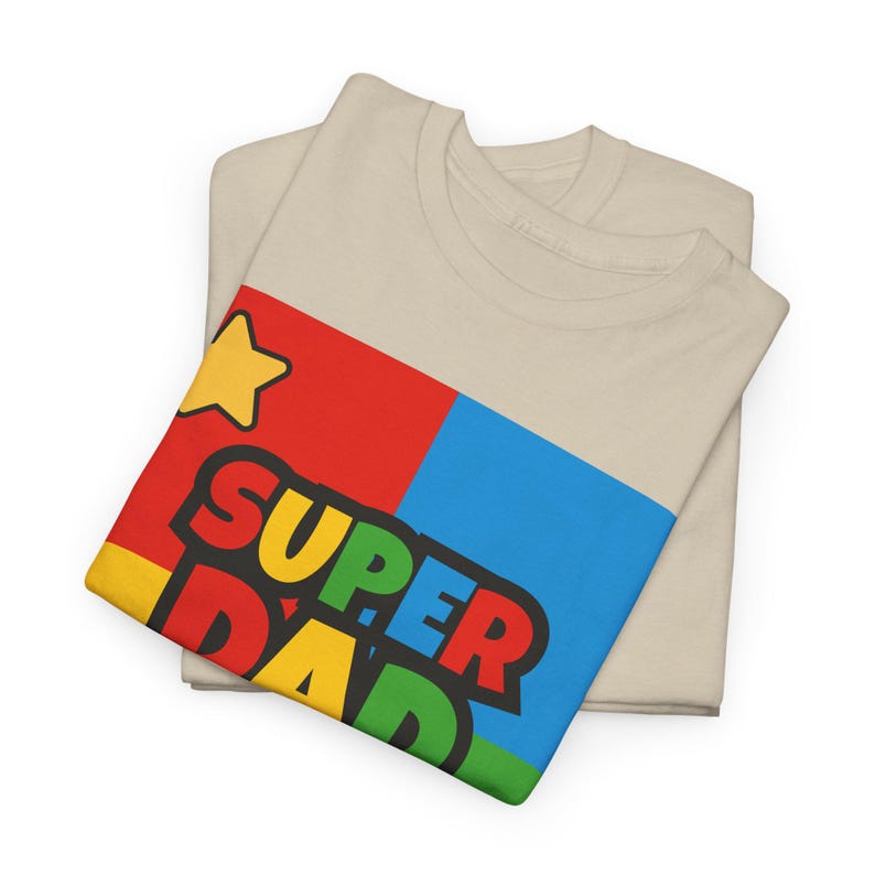 May include: Two beige t-shirts are folded, with one on top featuring a colorful graphic. The graphic includes a yellow star and the words "SUPER DAD" in bold, multi-colored letters against a red, blue, and yellow background. The shirts are made of a soft material.