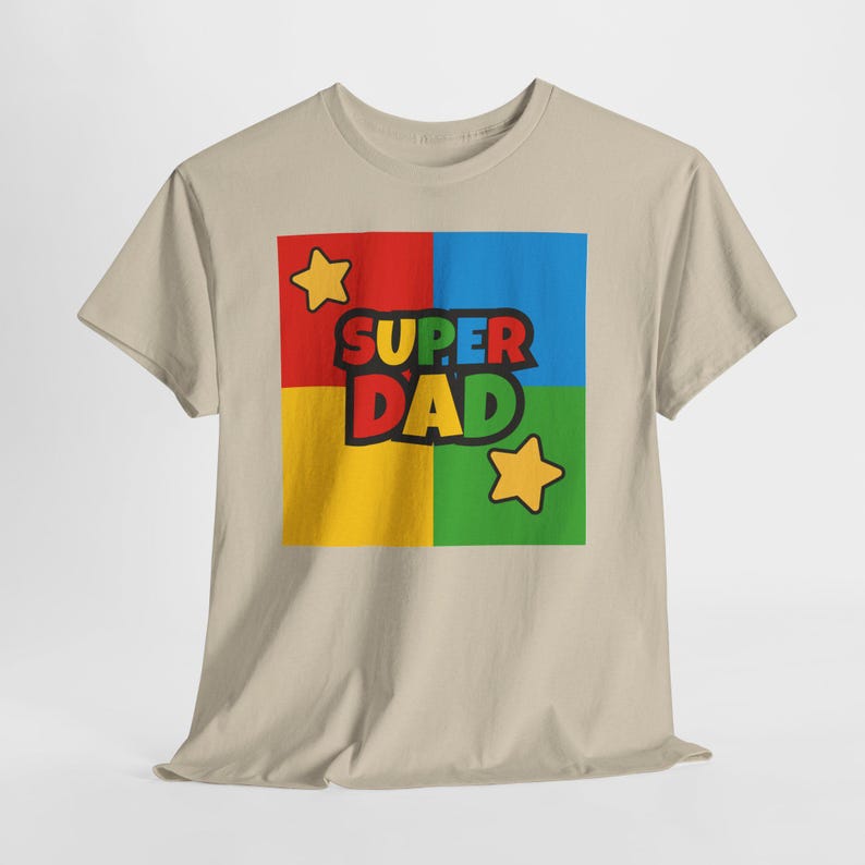 May include: A beige t-shirt featuring a colorful graphic that reads "SUPER DAD." The graphic is divided into four squares of red, blue, yellow, and green, with yellow stars in the corners. The text is in black with a yellow outline.