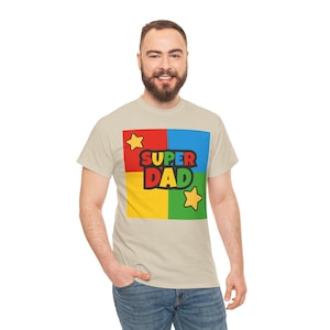 May include: Beige t-shirt with a colorful graphic that says "SUPER DAD." The graphic is in a square divided into four sections: red, blue, yellow, and green. Two yellow stars are on the graphic.