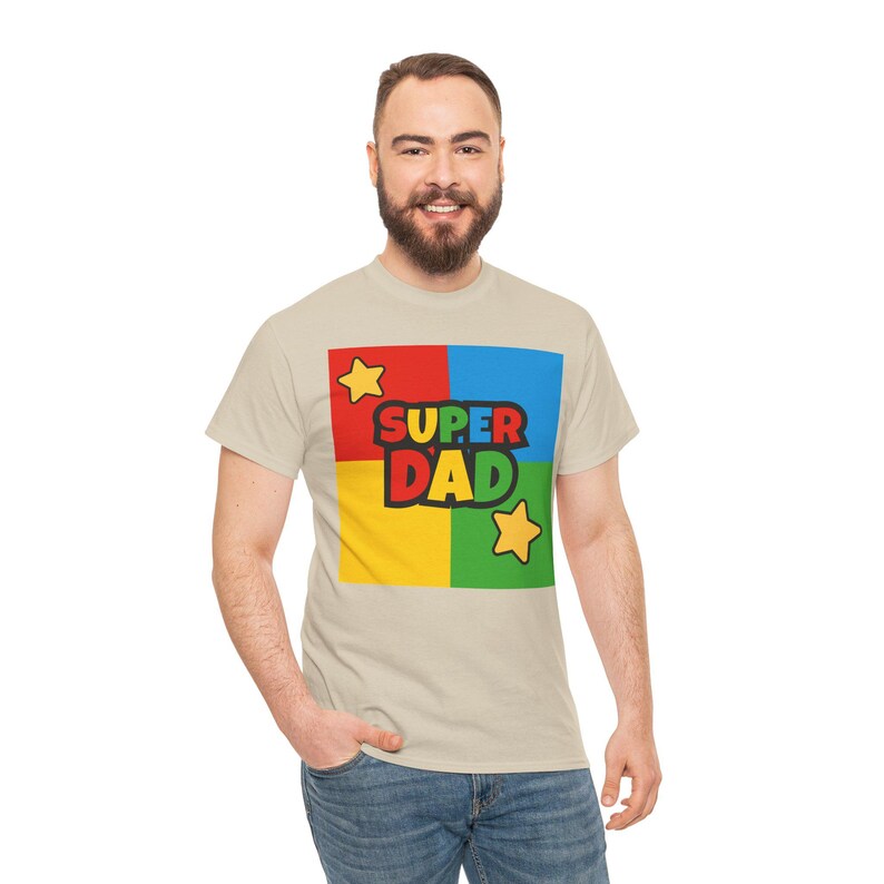 May include: Beige t-shirt with a colorful graphic that says "SUPER DAD." The graphic is in a square divided into four sections: red, blue, yellow, and green. Two yellow stars are on the graphic.