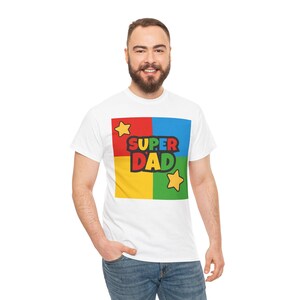 May include: White t-shirt with a colorful graphic that says "SUPER DAD". The graphic is divided into four squares of red, blue, yellow, and green, with yellow stars in the corners. A casual, everyday tee.