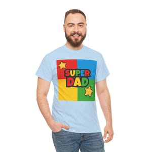 May include: Light blue t-shirt with a colorful graphic that says "SUPER DAD". The graphic is in a square divided into four sections: red, blue, yellow, and green. Two yellow stars are also included in the design.
