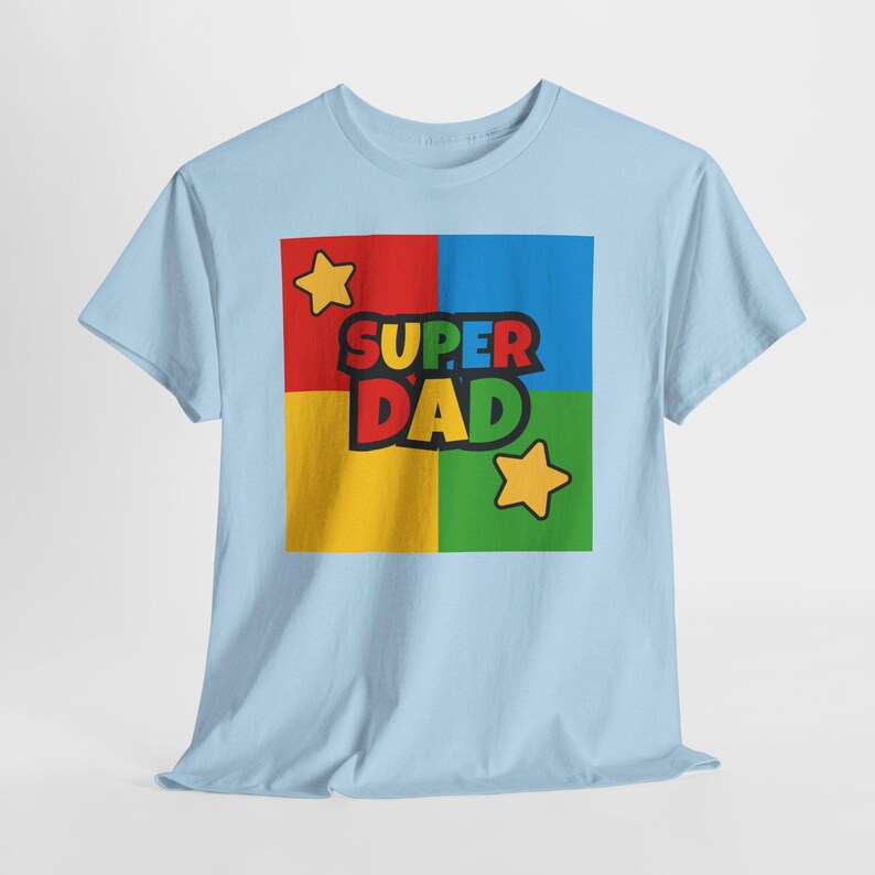 May include: Light blue t-shirt with a colorful graphic. The graphic features the words "SUPER DAD" in black and green, set against a square divided into red, blue, yellow, and green sections. Two yellow stars are also included.