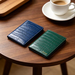 May include: Two rectangular passport holders with a crocodile skin texture. One holder is dark blue with the name Evelyn embossed on the front, and the other is dark green with the name Jesse embossed in gold lettering on the front surface.