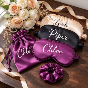 May include: Three satin sleep masks in champagne, black, and purple, each personalised with names Leah, Piper, and Chloe. A matching purple drawstring pouch labelled Chloe and a purple scrunchie are included, accompanied by a bouquet of peach roses.