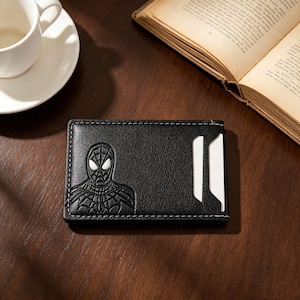 May include: A black leather card holder featuring an embossed Spider-Man character design on the left side and two vertical card slots on the right side with white stitching along the edges of the rectangular wallet.