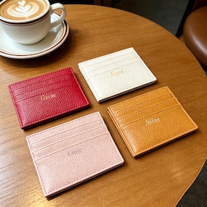 May include: Four leather cardholders in red, white, pink, and tan, each with a name embossed on the front. The cardholders have multiple card slots and are arranged on a wooden surface. A cup of coffee sits in the background.