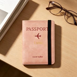 Könnte beinhalten: Ein hellrosa Reisepass-Etui mit einem schwarzen elastischen Verschlussband. Auf dem Einband steht das Wort PASSPORT in Dunkelrot, ein rotes Flugzeugsymbol, der Name Linda in Gold und der Text travel wallet in Dunkelrot.