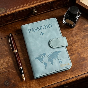 May include: A light blue passport holder with a matte finish, featuring the word PASSPORT, an airplane icon, a world map graphic, and the name Gavin embossed in gold. The cover includes a side strap closure and visible stitching along the edges.