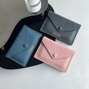 May include: Three envelope-style card holders in blue, dark grey, and light pink with a textured finish. Each features a matching snap button closure and a personalised name embossed in gold or silver: Grace, Orion, and Hana.