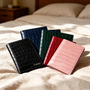 May include: A selection of passport holders in black, navy, green, red, and pink. Each has a crocodile skin effect. The black holder is personalized with the name "Lacey". The pink holder has the initials "TOH". The holders are arranged on a bed.