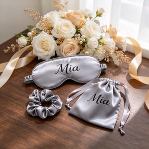 May include: A silver-gray satin sleep mask, a matching scrunchie, and a drawstring pouch, each featuring the name Mia in black script. The items are arranged together, with the sleep mask and pouch both clearly displaying the personalized text.