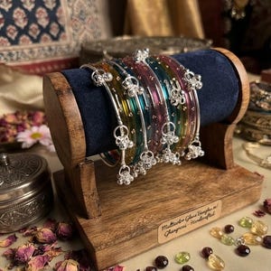 Peut inclure: Un ensemble de bracelets en verre translucide jaune, bleu sarcelle et magenta, ornés de petites breloques en métal argenté en forme de clochettes. Les bracelets sont présentés sur un support en bois avec un rouleau en velours bleu et une étiquette portant l'inscription Multicolor Glass Bangles / Ghungroos - Handcrafted.