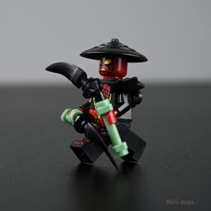 May include: A black and red minifigure with a black hat and a curved blade. The figure has a red head with yellow markings and is holding a green and black weapon. The figure is posed in a fighting stance.