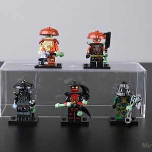 May include: Five Lego minifigures of ninja characters are displayed on a clear acrylic stand. The figures are in various colors, including orange, black, red, and green, and are equipped with weapons and armor. The background is a light gray.