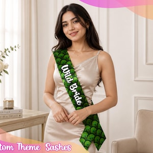May include: A sash featuring a green scale pattern and the white text Wild Bride. The sash is worn diagonally across the torso over a light-coloured sleeveless dress. The fabric displays a repeating rounded scale design in various shades of green.
