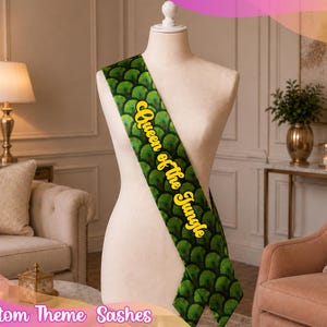 May include: A sash draped over a mannequin featuring a repeating green and black peacock feather pattern. The text "Queen of the Jungle" is printed in a bold, yellow, cursive font running diagonally across the center of the fabric sash.