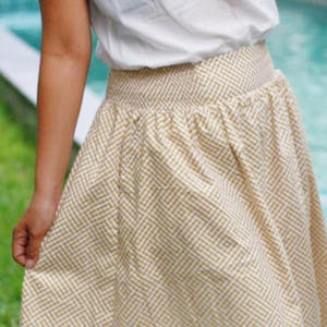 May include: A beige and white patterned skirt with a gathered waistband. The skirt has a geometric design and is knee-length. The person is wearing a white top. The skirt is made of a lightweight fabric.