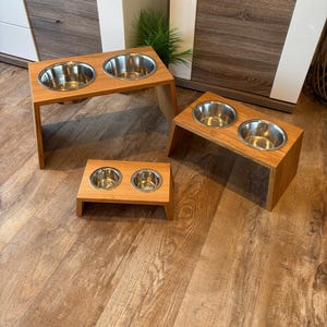 Puede incluir: Tres estaciones de alimentación para mascotas de madera en diferentes alturas, cada una con dos cuencos circulares de acero inoxidable. Los soportes están hechos de madera clara con un acabado liso para sostener los cuencos firmemente.
