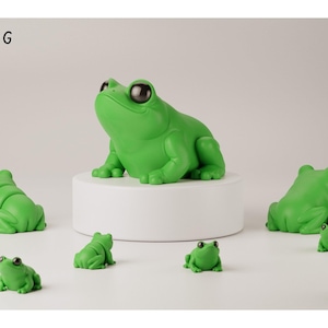 May include: A collection of bright green frog figurines in various sizes, featuring smooth surfaces and large, dark, glossy eyes. One large frog sits atop a white cylindrical pedestal, while several smaller frogs are positioned around it.