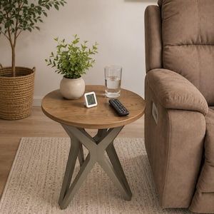 May include: A round side table with a light brown wood grain top and a gray X-shaped base. On the table sits a small potted green plant in a beige ceramic pot, a digital thermometer displaying 24 and 56, a clear glass of water, and a black remote.