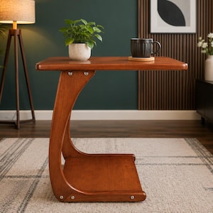 May include: A C-shaped side table crafted from warm-toned wood, featuring a flat rectangular top surface and a lower shelf connected by a curved support frame. The table is secured with visible metal hardware at the joints and has a smooth finish.