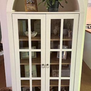 May include: A white arched cabinet with two glass-paned doors and a bottom drawer. The interior features wooden shelves displaying various decorative items, including a sign that reads love grows best in little houses just like this.