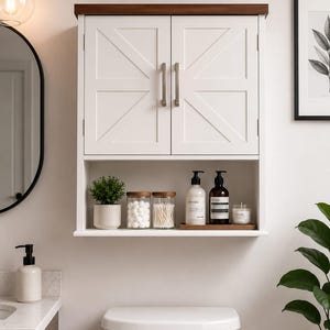 May include: A white wall-mounted bathroom cabinet with two doors featuring a diagonal cross-brace design, silver handles, and a dark wood top shelf. An open storage shelf below holds a small potted plant, two glass jars, and two pump bottles.