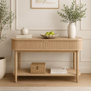 May include: A light wood console table with a fluted front, two small cabinet doors, and a lower shelf. The table holds a white vase with green branches, a scalloped bowl with green apples, a small round vase, and a book titled KINFOLK.