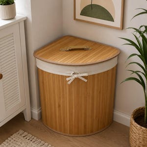 May include: A corner-shaped laundry hamper constructed from vertical bamboo slats with a matching lid featuring a tan handle. The top edge is finished with a beige fabric band secured by a tied bow at the front center, providing a structured look.