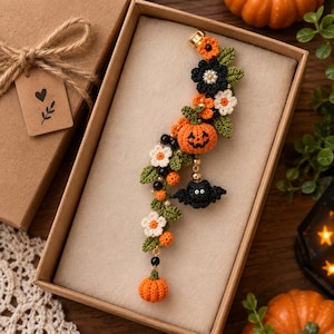 May include: A decorative crochet ear cuff featuring a curved vine design with orange pumpkins, black and white flowers, green leaves, and a small black bat charm. The piece is accented with small black and white beads and gold-toned metal hardware.