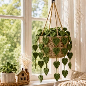May include: A beige crocheted hanging planter basket containing a trailing plant with numerous green crocheted heart-shaped leaves. The planter is suspended by three braided beige cords attached to a central ring at the top of the arrangement.