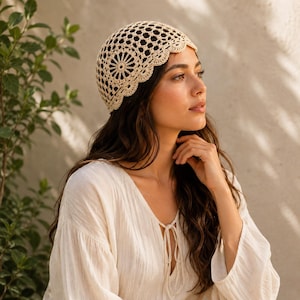 May include: A cream-colored crochet cap featuring an intricate openwork pattern with a circular motif at the side and a scalloped edge. The headwear is crafted from fine yarn, showcasing a delicate, detailed texture throughout the entire design.