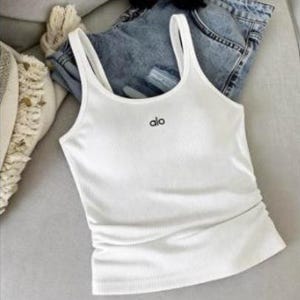 May include: A white ribbed tank top with the word "alo" in black lettering. The tank top has thin straps and is paired with blue denim shorts and black sunglasses. The items are arranged on a light gray surface.