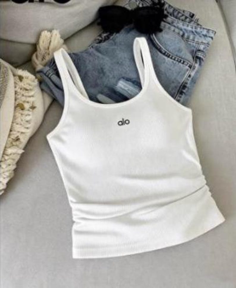 May include: A white ribbed tank top with the word "alo" in black lettering. The tank top has thin straps and is paired with blue denim shorts and black sunglasses. The items are arranged on a light gray surface.