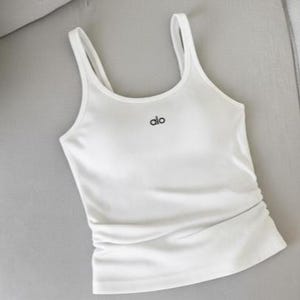 May include: White tank top with thin straps and the word "alo" in black script. The top has a fitted silhouette and a gathered detail at the waist. The fabric appears smooth and slightly shiny.