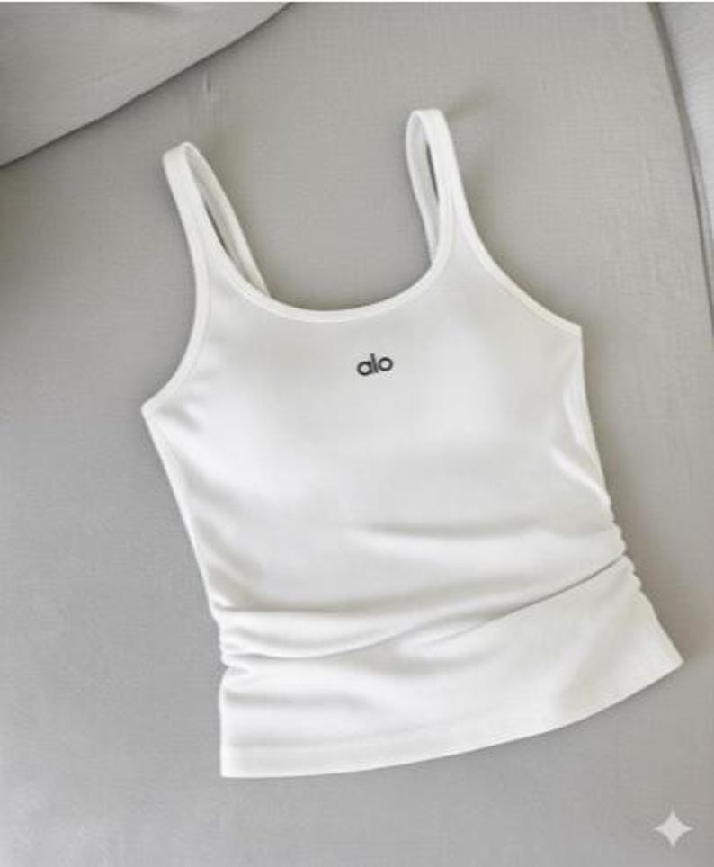 May include: White tank top with thin straps and the word "alo" in black script. The top has a fitted silhouette and a gathered detail at the waist. The fabric appears smooth and slightly shiny.