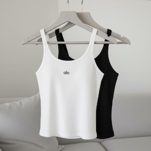 May include: Two ribbed tank tops, one white and one black, hang on gray hangers. The white tank top has the word "alo" printed in black on the front. The tank tops are displayed against a neutral background.