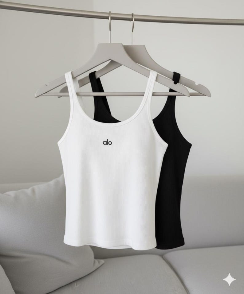 May include: Two ribbed tank tops, one white and one black, hang on gray hangers. The white tank top has the word "alo" printed in black on the front. The tank tops are displayed against a neutral background.