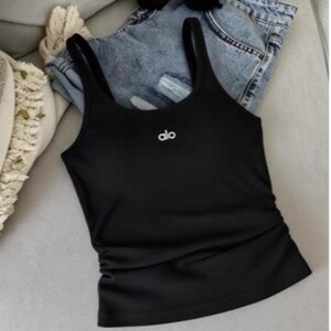 May include: A black tank top with thin straps and the brand name "alo" in white lettering. The tank top is paired with blue denim shorts and sunglasses. The top has a ruched detail on the side.