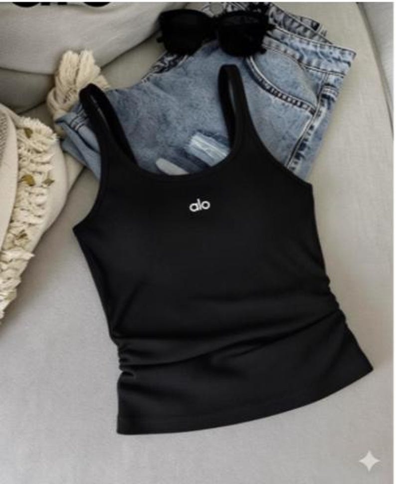 May include: A black tank top with thin straps and the brand name "alo" in white lettering. The tank top is paired with blue denim shorts and sunglasses. The top has a ruched detail on the side.