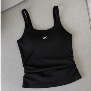 May include: Black ribbed tank top with thin straps. The word "alo" is printed in white on the chest. The tank top has a fitted silhouette and a gathered detail on the side.