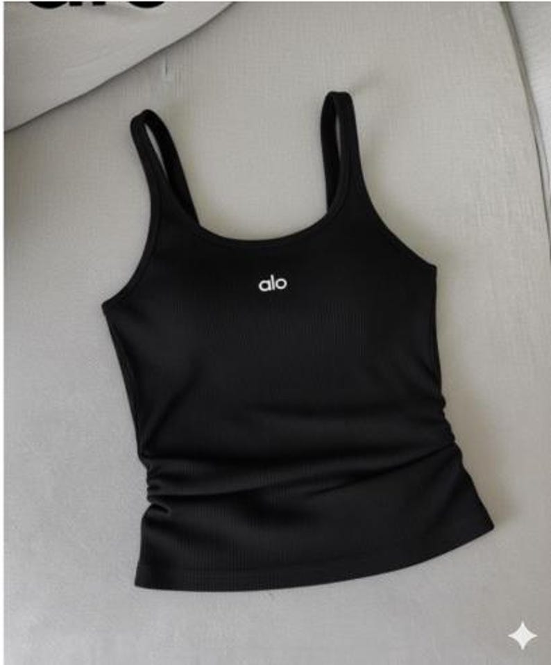 May include: Black ribbed tank top with thin straps. The word "alo" is printed in white on the chest. The tank top has a fitted silhouette and a gathered detail on the side.