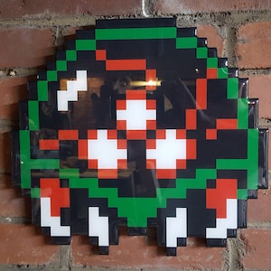 May include: A black, green, red, and white pixelated wall art piece depicting a video game character. The character has three eyes and a mouth.