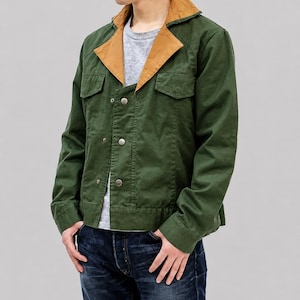 Könnte beinhalten: Eine dunkelgrüne Jacke mit kontrastierendem beigefarbenem Kragen und Revers, langen Ärmeln, zwei Brusttaschen mit Klappen und einer Knopfleiste mit drei sichtbaren Metallknöpfen, getragen über einem grauen T-Shirt und einer dunkelblauen Jeans.