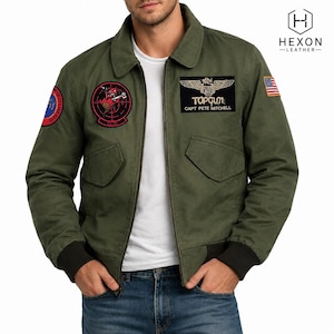 May include: An olive green bomber jacket worn over a white t-shirt, featuring a black Top Gun patch with gold wings, a circular red and black target patch, a circular blue and red patch, and a small American flag patch on the left sleeve.