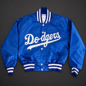 Può includere: Una giacca varsity in raso blu reale con la scritta Dodgers in bianco sul petto. La giacca presenta una chiusura con bottoni a pressione, un colletto a coste e polsini a coste con due strisce bianche. Il tessuto ha una finitura liscia e lucida.