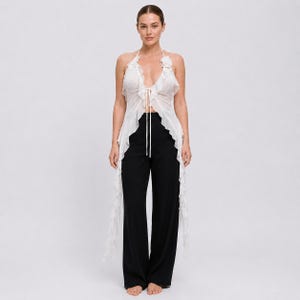 May include: A white sleeveless halter top featuring a deep V-neckline, a front tie closure, and long ruffled edges that cascade down the sides. The top is paired with solid black wide-leg trousers that feature a clean, straight-cut silhouette.