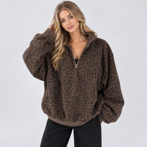Op de afbeelding: Een bruine en donkerbruine fleece trui met luipaardprint, voorzien van een opstaande kraag, een korte rits, lange mouwen met geribbelde boorden en een geribbelde zoom. Het kledingstuk heeft een relaxte pasvorm en een zachte textuur.
