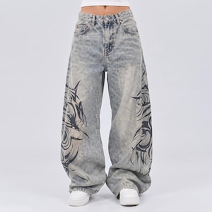 May include: Light blue acid-wash denim jeans with a wide-leg silhouette. The trousers feature a high-waisted fit with a button and zip fastening, five pockets, and large, dark, swirling tribal-inspired graphic prints on both lower trouser legs.